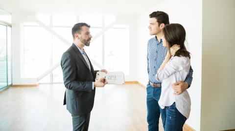 Un professionnel, un rapport à la main, donne des explications à un jeune couple debout face à lui dans un appartement. 