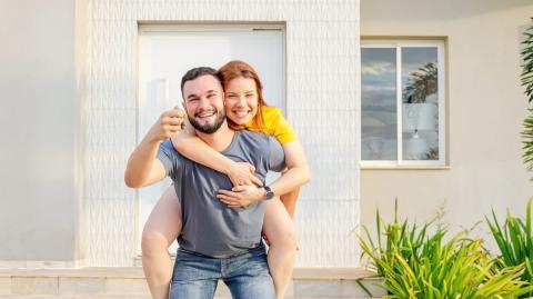 Un jeune homme souriant, qui porte sa compagne à califourchon sur son dos, montre fièrement les clés de leur nouvelle maison