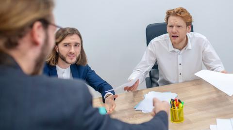 Deux hommes assis devant un bureau font face à un homme énervé qui leur tend un document