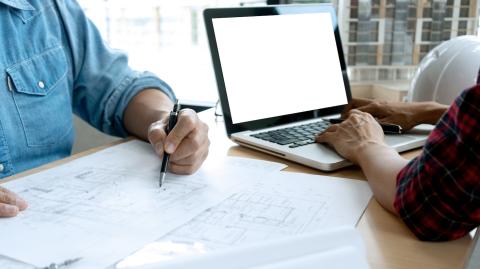 2 personnes travaillent face à face sur un bureau commun, la femme travaille sur son pc, l'homme, sur le plan d'un bâtiment