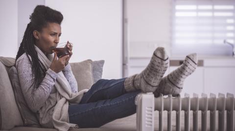 Une femme assise sur son canapé essaie de se réchauffer, en posant ses pieds couverts de grosses chaussettes épaisses contre un radiateur et en buvant une boisson chaude