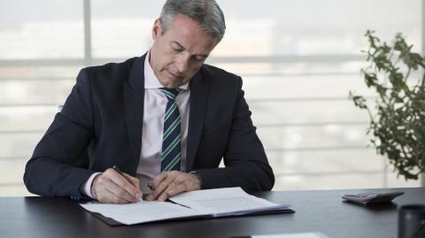 Un homme d'âge mûr en costume signe des documents, assis à son bureau