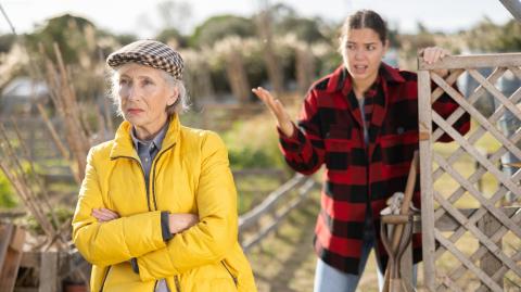 Une jeune femme, appuyée contre la barrière de son terrain, a un différend avec une voisine plus âgée qui s'éloigne en lui tournant le dos