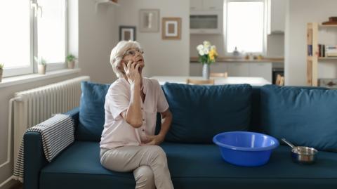 Une femme d'âge mûr, assise sur son canapé, est en communication téléphonique au sujet d'un dégât des eaux dans son salon