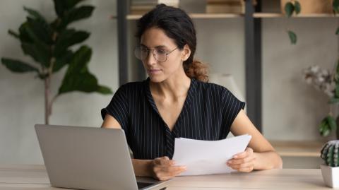 Une femme portant des lunettes et les cheveux attachés consulte des documents sur son pc portable, un document papier dans les mains