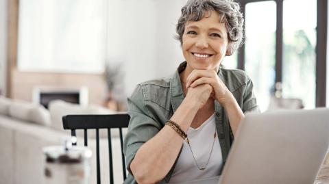 Une femme aux cheveux courts gris, assise devant son pc portable, affiche un sourire satisfait