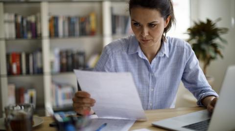 Une femme règle des questions administratives, assise devant son ordinateur portable, un document dans les mains