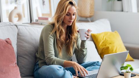 Une jeune femme aux longs cheveux blonds effectue des démarches sur son pc portable, assise en tailleur sur son canapé