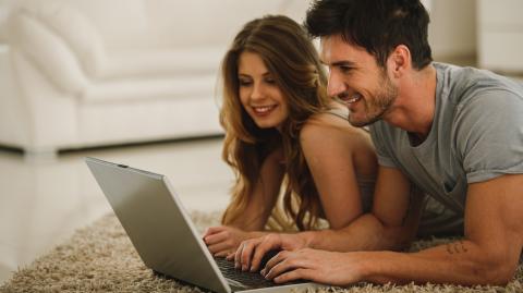 Un jeune homme et une jeune femme, à plat ventre sur le tapis de leur salon, font une recherche sur un pc portable