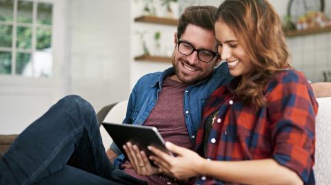 Un couple souriant, installé sur un canapé, réalise une étude sur une tablette