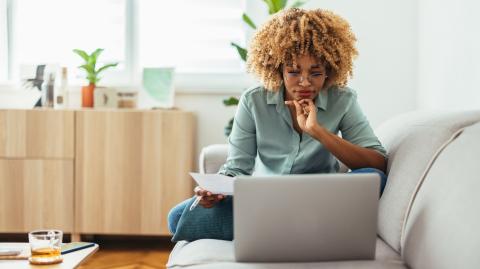 Une jeune femme aux cheveux crépus, assise en tailleur sur son canapé, réalise une étude sur son pc portable
