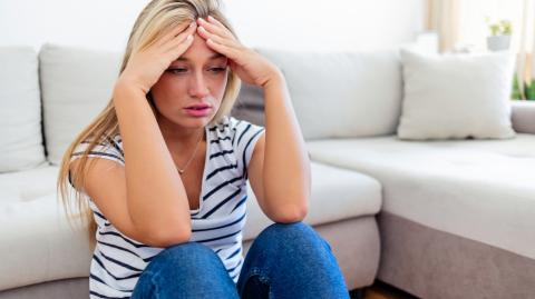 Une jeune femme aux longs cheveux blonds est assise par terre près de son canapé et tient sa tête entre ses mains, perturbée par un événement récent