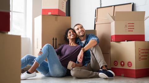 Un jeune couple est assis par terre enlacé, en admirant son nouveau logement rempli de cartons de déménagement