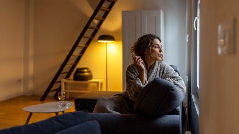 Une femme, assise sur son canapé, regarde par la fenêtre en fumant une cigarette