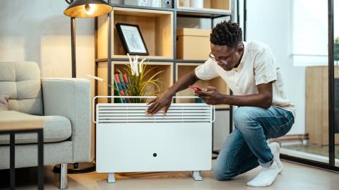 Un homme règle la consommation de son radiateur électrique à l'aide de son smartphone, agenouillé devant l'appareil dans son salon