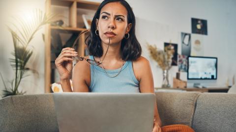 Une jeune femme réfléchit dans son salon devant son ordinateur, une paire de lunettes à la main