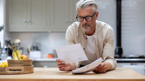 Un homme aux cheveux blancs lit un courrier, en appui sur le plan de travail de sa cuisine