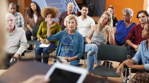 Une femme assise dans l'assemblée lève la main pour parler lors d'une assemblée générale de copropriété