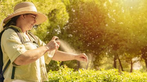 Une dame portant un chapeau vaporise un spray anti-moustiques sur son avant-bras, en extérieur