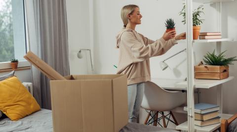 Une étudiante défait ses cartons et pose une plante sur une étagère pour la décorer