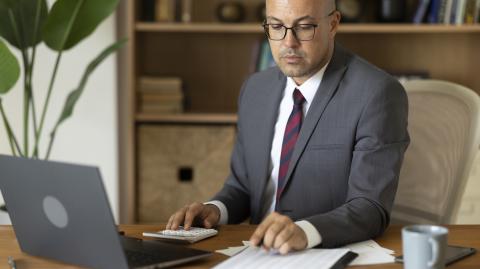Un homme en costume travaille sur son pc tout en faisant des calculs, assis à son bureau