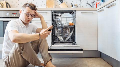 Un homme est assis sur le sol de sa cuisine devant son lave-vaisselle ouvert, son téléphone à la main