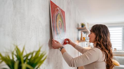 Une jeune femme aux cheveux longs ondulés accroche un cadre dans son salon, sans percer les murs