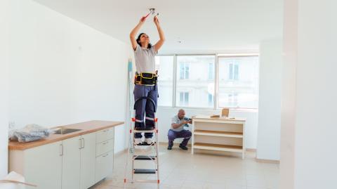 Une électricienne procède à l'installation d'un luminaire dans une cuisine, debout sur un escabeau