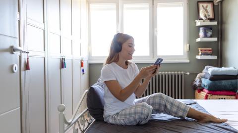 Une jeune femme blonde en pyjama regarde son téléphone, assise sur son lit avec un casque audio sur les oreilles