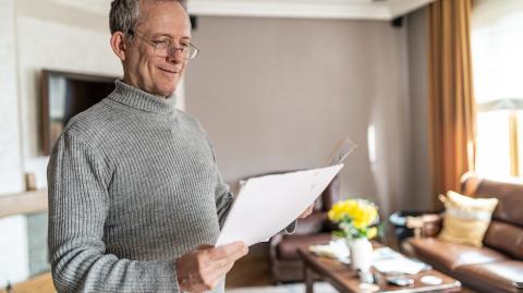 Un homme d'âge mûr examine un document debout dans son salon, avec le sourire