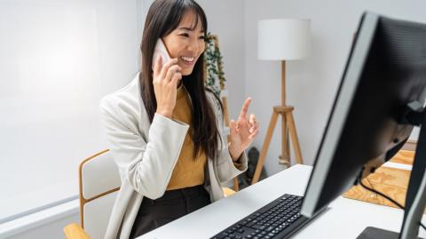 Une femme aux cheveux longs est en pleine conversation téléphonique, assise devant son ordinateur 
