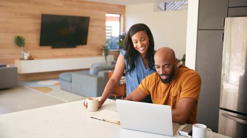 Un couple souriant fait des recherches sur ordinateur sur la table de sa cuisine ouverte sur le salon