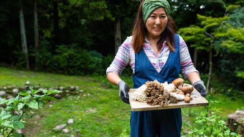 Une femme marche dans son jardin, en portant une planche en bois avec les champignons qu'elle vient de ramasser