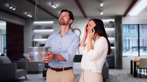 Un homme tient une bassine pour récupérer l'eau qui coule du plafond dans son salon tandis que sa femme à côté de lui téléphone