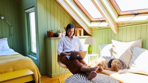 Une jeune femme se détend assise avec son ordinateur sur les genoux dans son studio sous les toits
