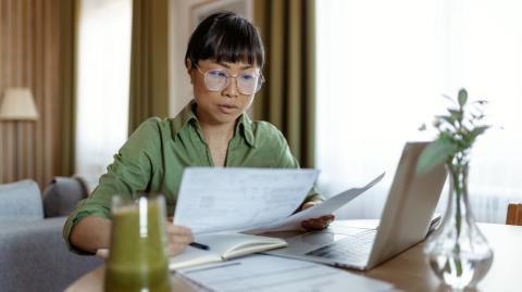 Une femme assise devant son pc portable dans son salon étudie des documents au format papier