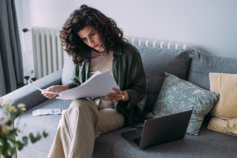 Femme travaillant sur son canapé gris, lisant ses documents, avec son ordinateur posé non loin. 