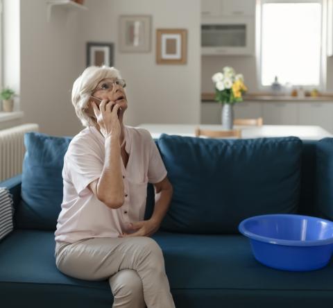 Une femme d'âge mûr, assise sur son canapé, est en communication téléphonique au sujet d'un dégât des eaux dans son salon