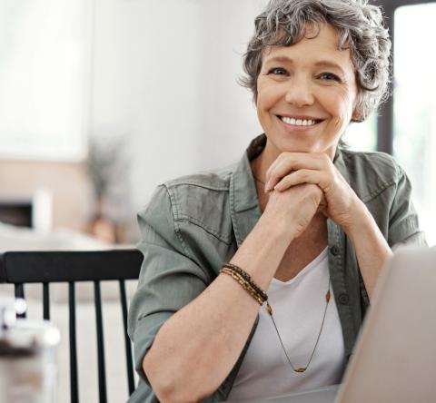 Une femme aux cheveux courts gris, assise devant son pc portable, affiche un sourire satisfait 