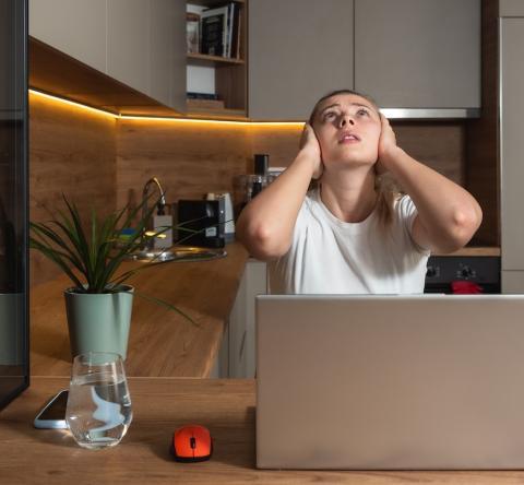 Une jeune femme travaillant sur son pc portable se bouche les oreilles en regardant le plafond de son appartement