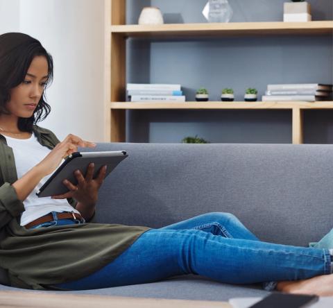 Une jeune femme d'origine asiatique, assise avec les jambes allongées sur son canapé, est concentrée sur le contenu de sa tablette numérique