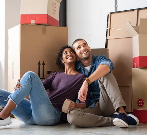 Un jeune couple est assis par terre enlacé, en admirant son nouveau logement rempli de cartons de déménagement