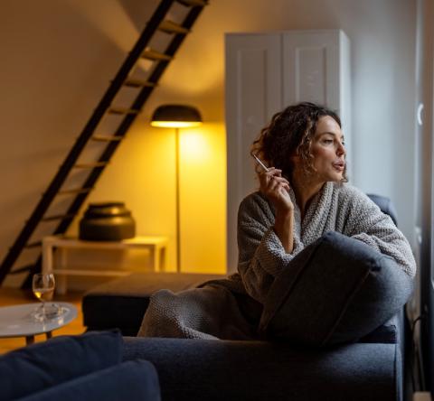 Une femme, assise sur son canapé, regarde par la fenêtre en fumant une cigarette