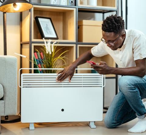 Un homme règle la consommation de son radiateur électrique à l'aide de son smartphone, agenouillé devant l'appareil dans son salon