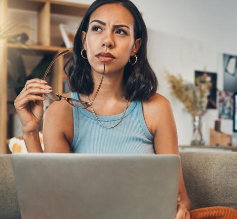Une jeune femme réfléchit dans son salon devant son ordinateur, une paire de lunettes à la main