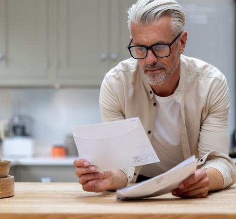 Un homme aux cheveux blancs lit un courrier, en appui sur le plan de travail de sa cuisine