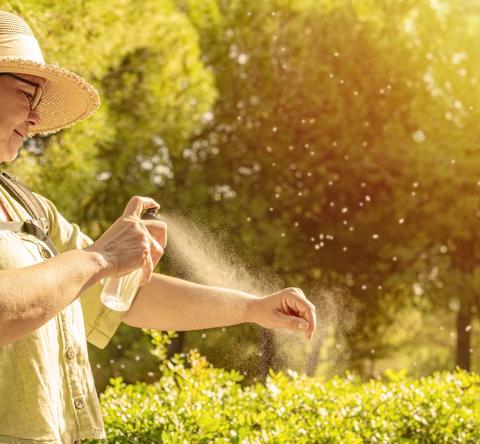Une dame portant un chapeau vaporise un spray anti-moustiques sur son avant-bras, en extérieur