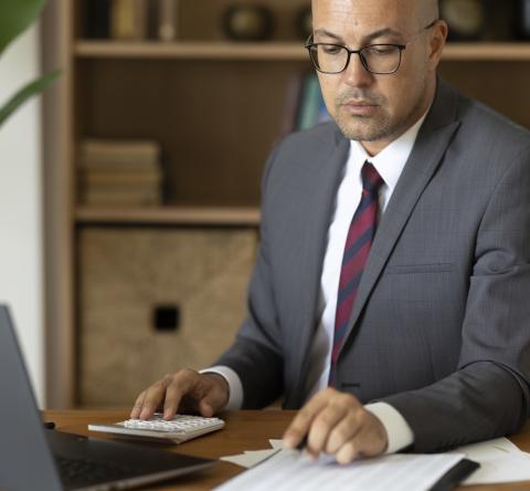Un homme en costume travaille sur son pc tout en faisant des calculs, assis à son bureau