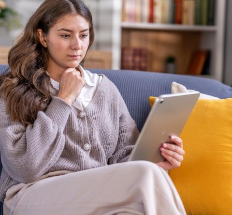 Une jeune femme étudie des documents sur une tablette, assise sur son canapé