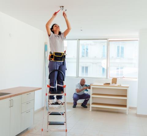 Une électricienne procède à l'installation d'un luminaire dans une cuisine, debout sur un escabeau
